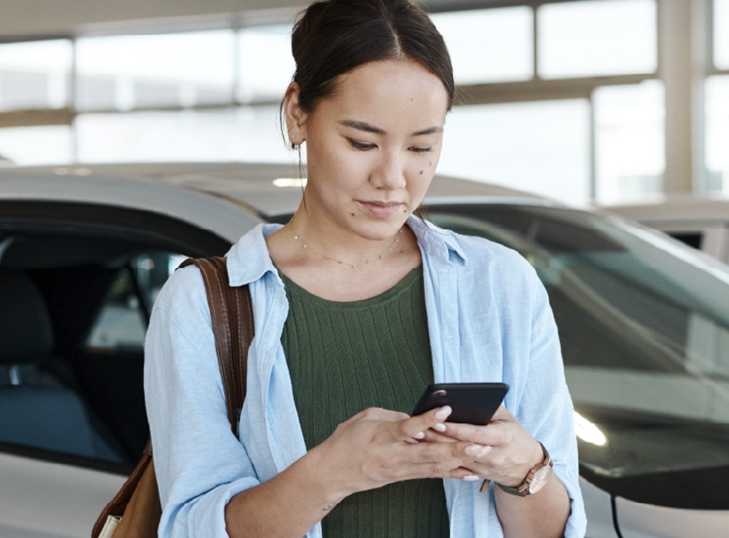 A woman stands next to a car in a showroom, focused on her phone while wearing a green top, blue shirt, and carrying a brown bag.